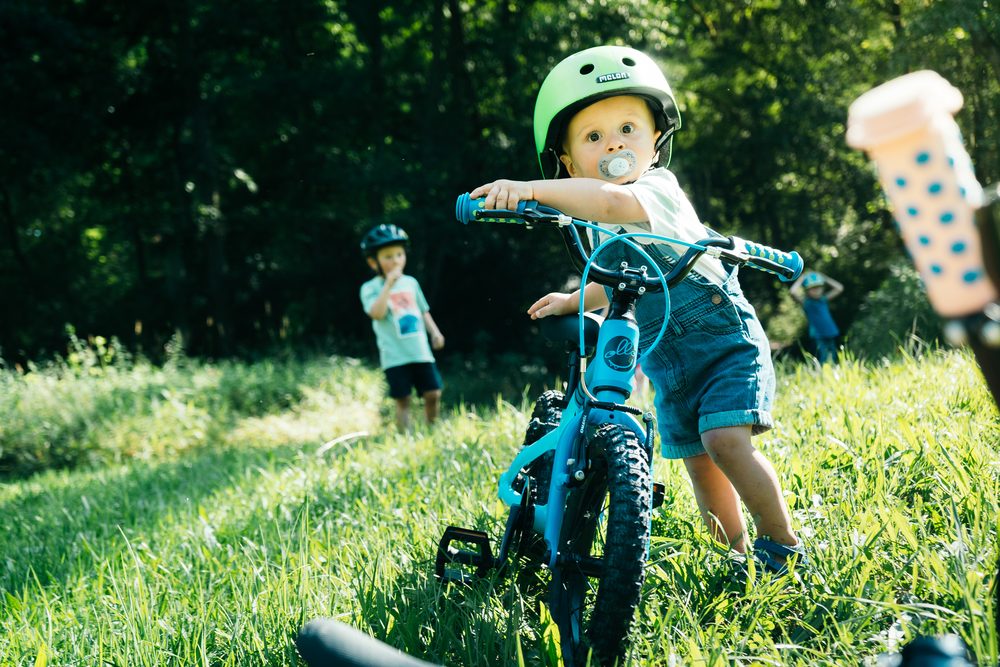 Kleinkind mit grünem Helm schiebt ein türkisfarbenes Ollo-Kinderfahrrad über eine sonnige Wiese