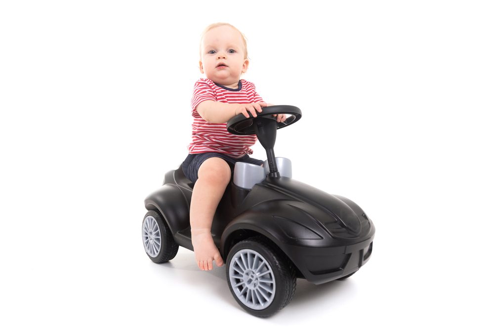 Kleinkind sitzt am Lenkrad eines schwarzen Rutschautos, freigestellt vor weißem Hintergrund